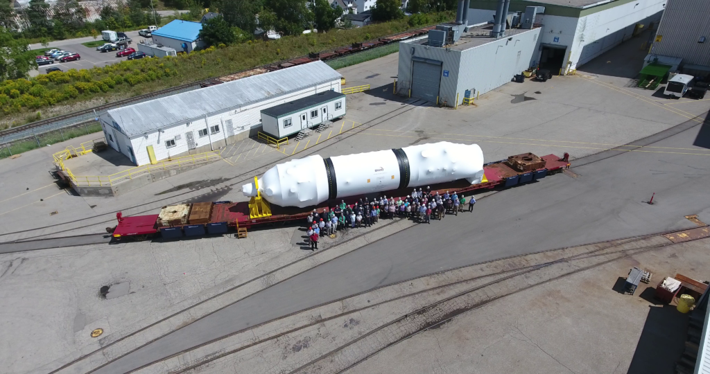 A BWXT steam generator ready for delivery and wrapped in white shipping materials at BWXT Cambridge, Ontario with a group of proud employees standing in front.